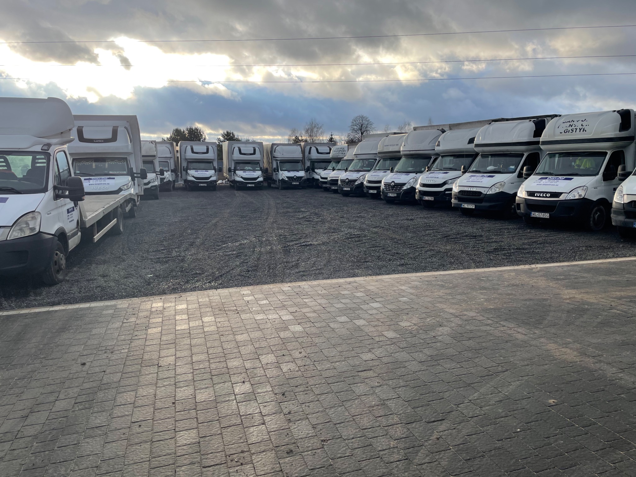 Flota białych samochodów dostawczych Iveco z zabudową kontenerową, zaparkowanych rzędami na żwirowym placu, w tle pochmurne niebo.