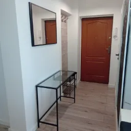 Wnętrze przedpokoju po remoncie: drewnopodobne panele podłogowe, białe ściany z czarnym lustrem i konsolą z metalową podstawą i szklanym blatem, brązowe drzwi wejściowe.