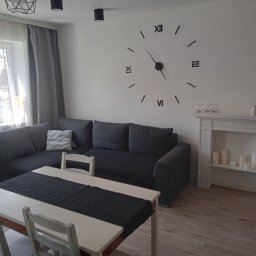 Michał Tessmer - Jasne wnętrze pokoju dziennego z szarą sofą narożną, białym stołem z ciemnym bieżnikiem, dekoracyjnym kominkiem z świecami i dużym zegarem ściennym w stylu rzymskim.