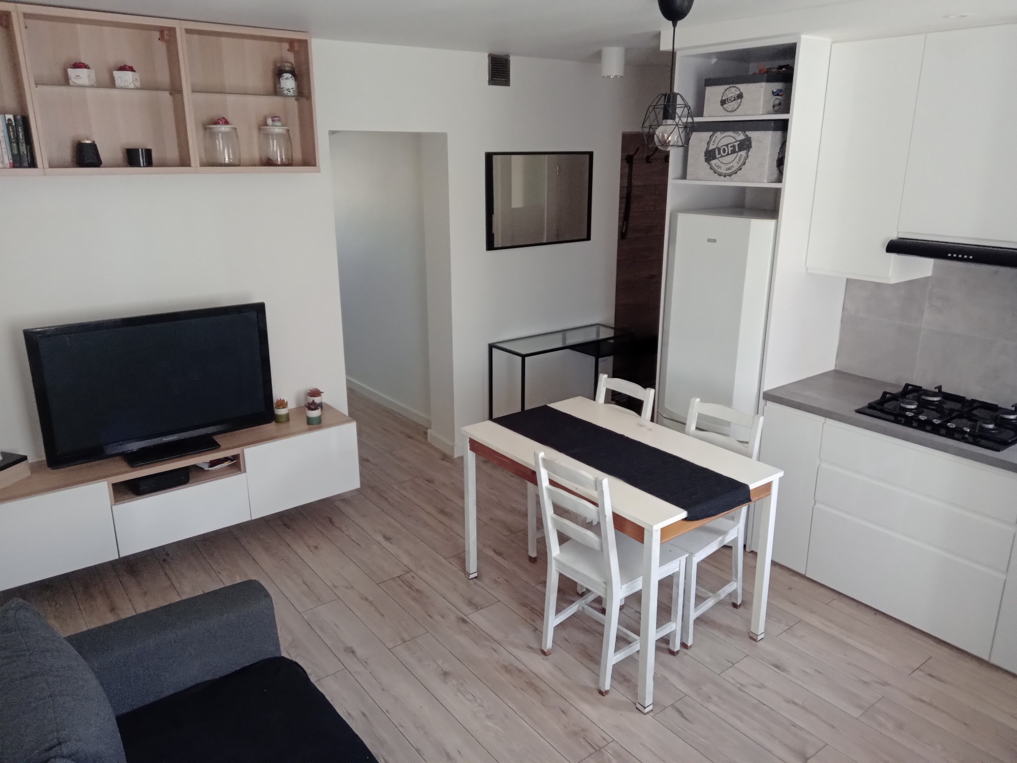 Jasne, przestronne wnętrze mieszkania z drewnianą podłogą, białymi meblami kuchennymi, stołem z krzesłami i telewizorem na komodzie.