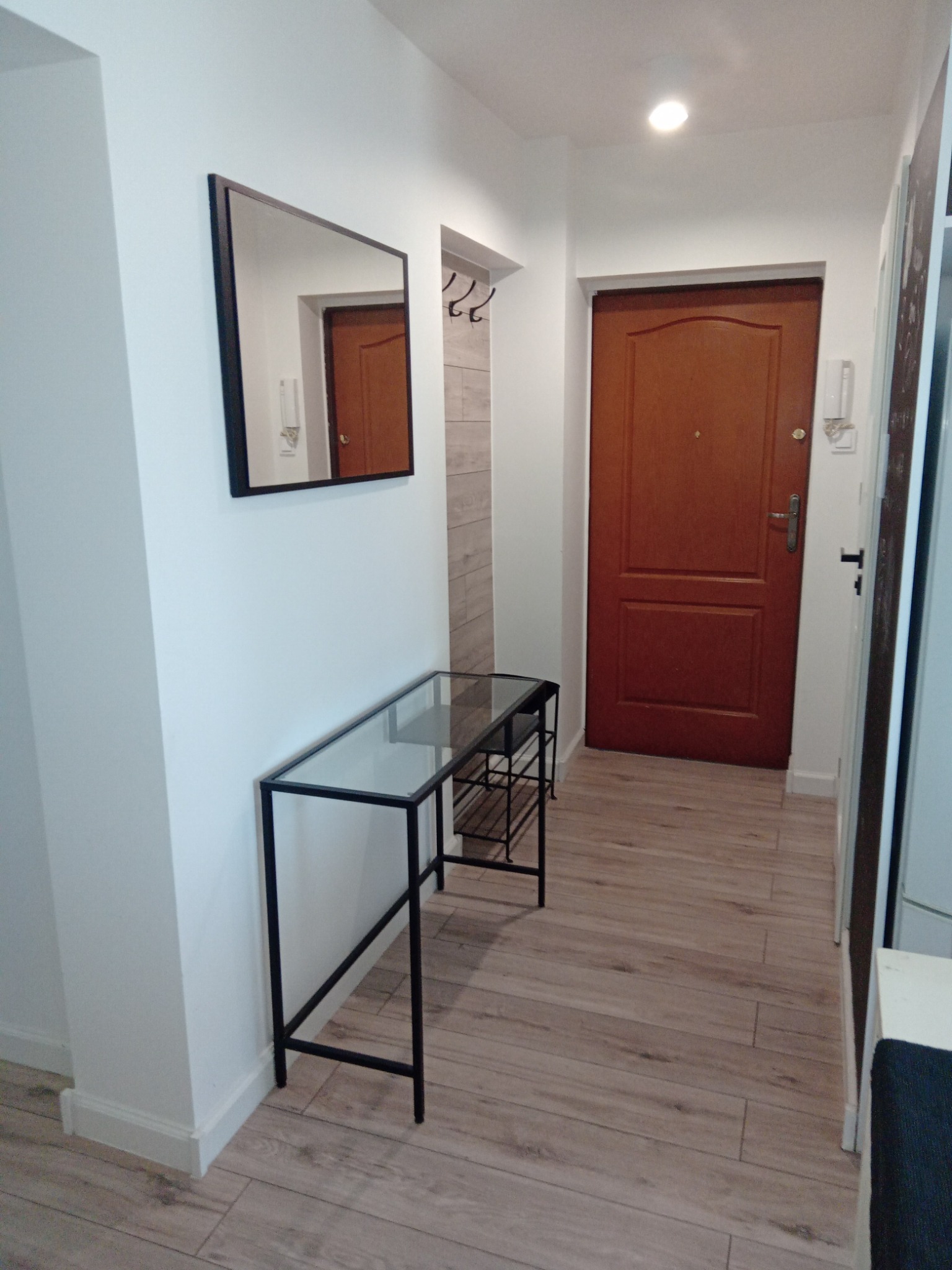 Wnętrze przedpokoju po remoncie: drewnopodobne panele podłogowe, białe ściany z czarnym lustrem i konsolą z metalową podstawą i szklanym blatem, brązowe drzwi wejściowe.