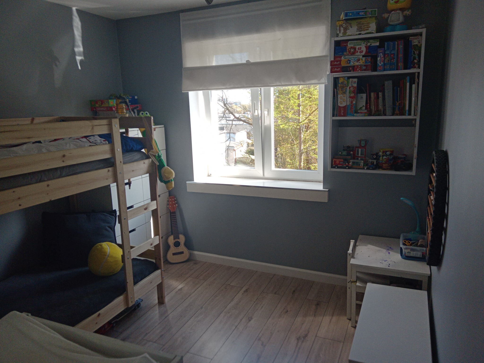 Pokój dziecięcy z drewnianym łóżkiem piętrowym, zasłoniętym oknem, regałem z książkami i zabawkami oraz drewnianą podłogą.