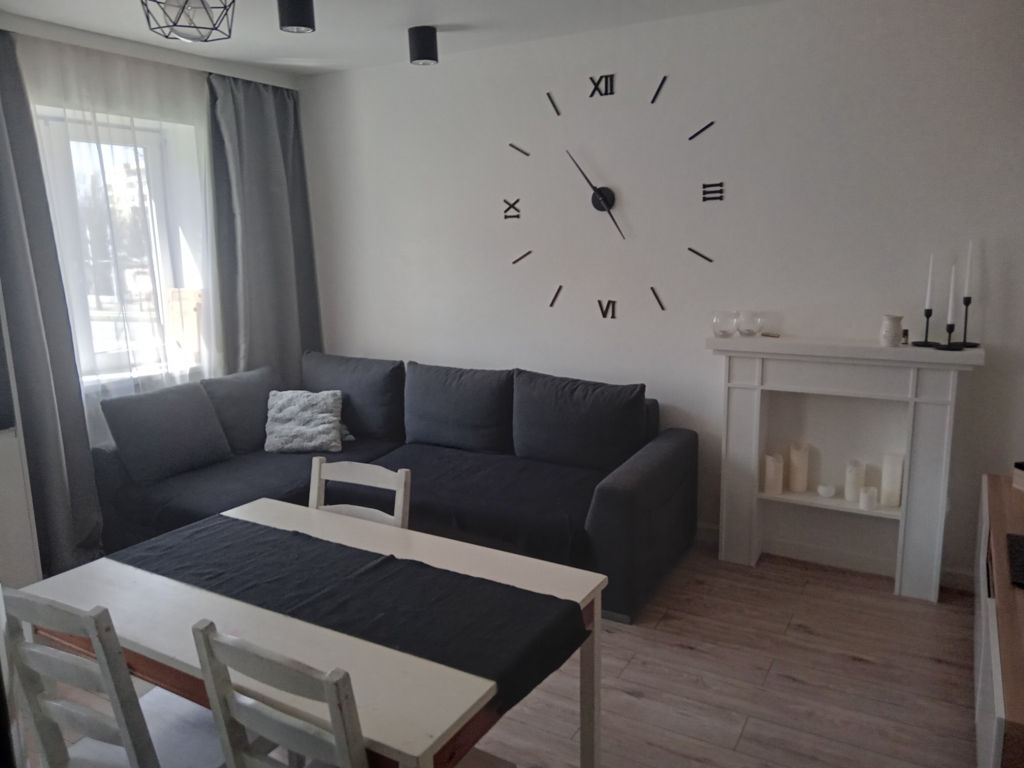 Jasne wnętrze pokoju dziennego z szarą sofą narożną, białym stołem z ciemnym bieżnikiem, dekoracyjnym kominkiem z świecami i dużym zegarem ściennym w stylu rzymskim.