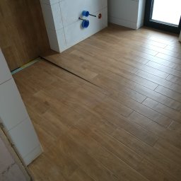 DP FENSTERTECHNIK - Ułożona podłoga z płytek imitujących drewno w łazience, widoczne białe płytki na ścianie z geometrycznym wzorem oraz niebieskie rury instalacji wodnej.