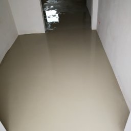 DP FENSTERTECHNIK - Świeżo wylana, gładka wylewka samopoziomująca na podłodze w trakcie remontu mieszkania, widoczna w perspektywie korytarza z białymi ścianami i odblaskami światła.