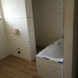 DP FENSTERTECHNIK - Wnętrze łazienki w trakcie wykańczania: białe, prostokątne płytki na ścianach, drewnopodobna podłoga, wanna zabezpieczona folią, widoczne instalacje hydrauliczne przed montażem armatury.