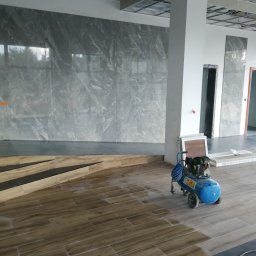 DP FENSTERTECHNIK - Wnętrze w trakcie prac wykończeniowych: ułożone płytki imitujące drewno na podłodze, szare marmurowe płytki na ścianie, widoczny kompresor i niezabudowany sufit podwieszany.
