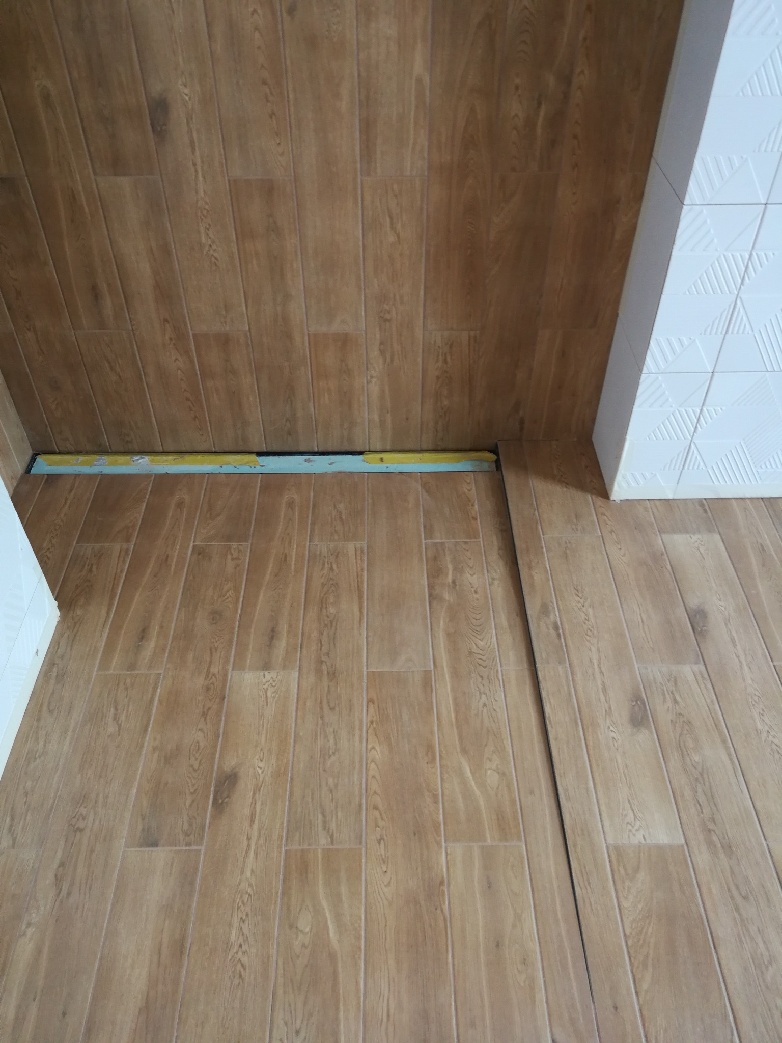 Kąt widzenia na nową podłogę z paneli imitujących drewno, ułożoną w małym pomieszczeniu; widoczny fragment białej ściany z dekoracyjnymi płytkami oraz fragment listwy przypodłogowej z resztkami...