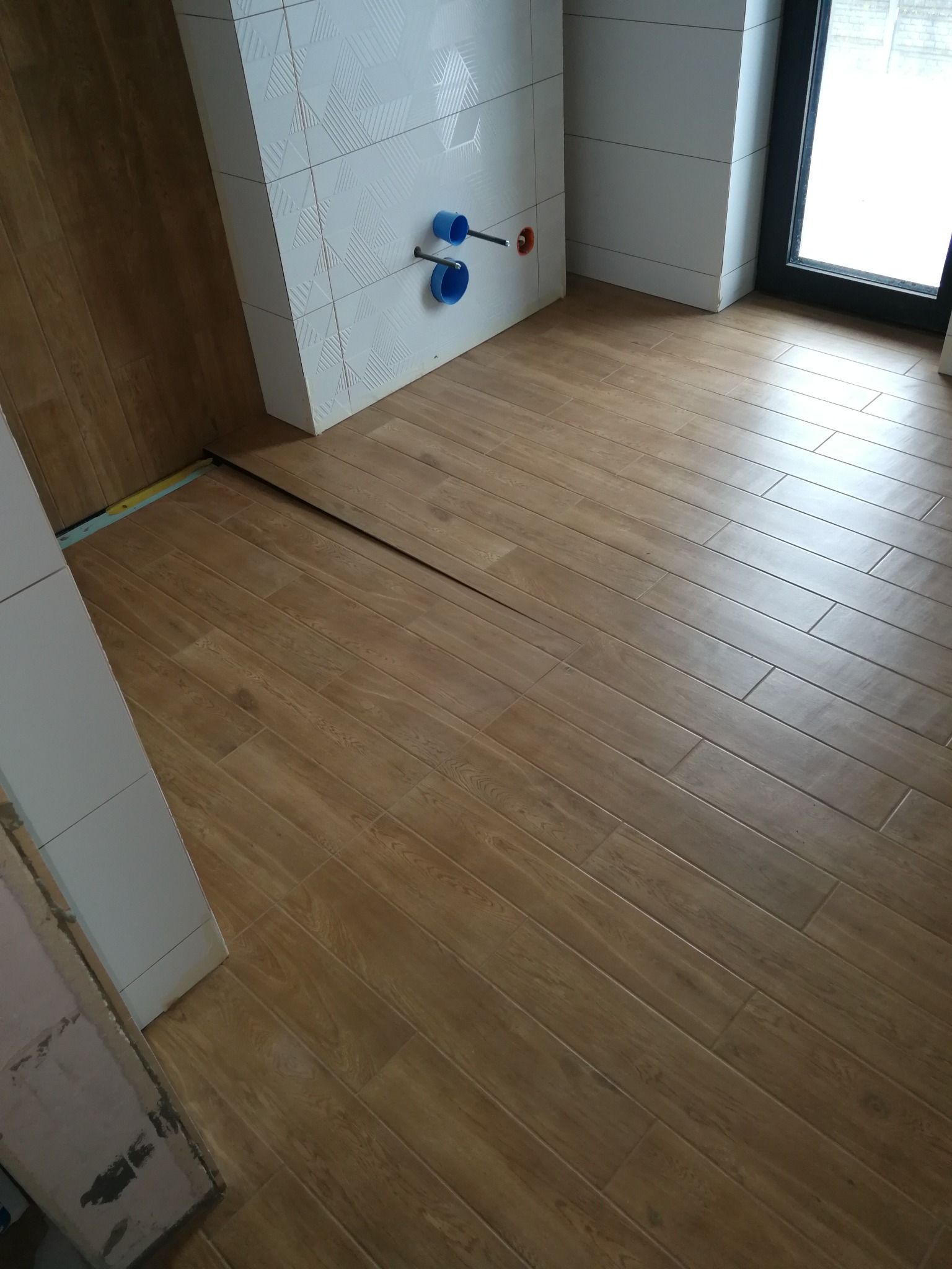 Ułożona podłoga z płytek imitujących drewno w łazience, widoczne białe płytki na ścianie z geometrycznym wzorem oraz niebieskie rury instalacji wodnej.