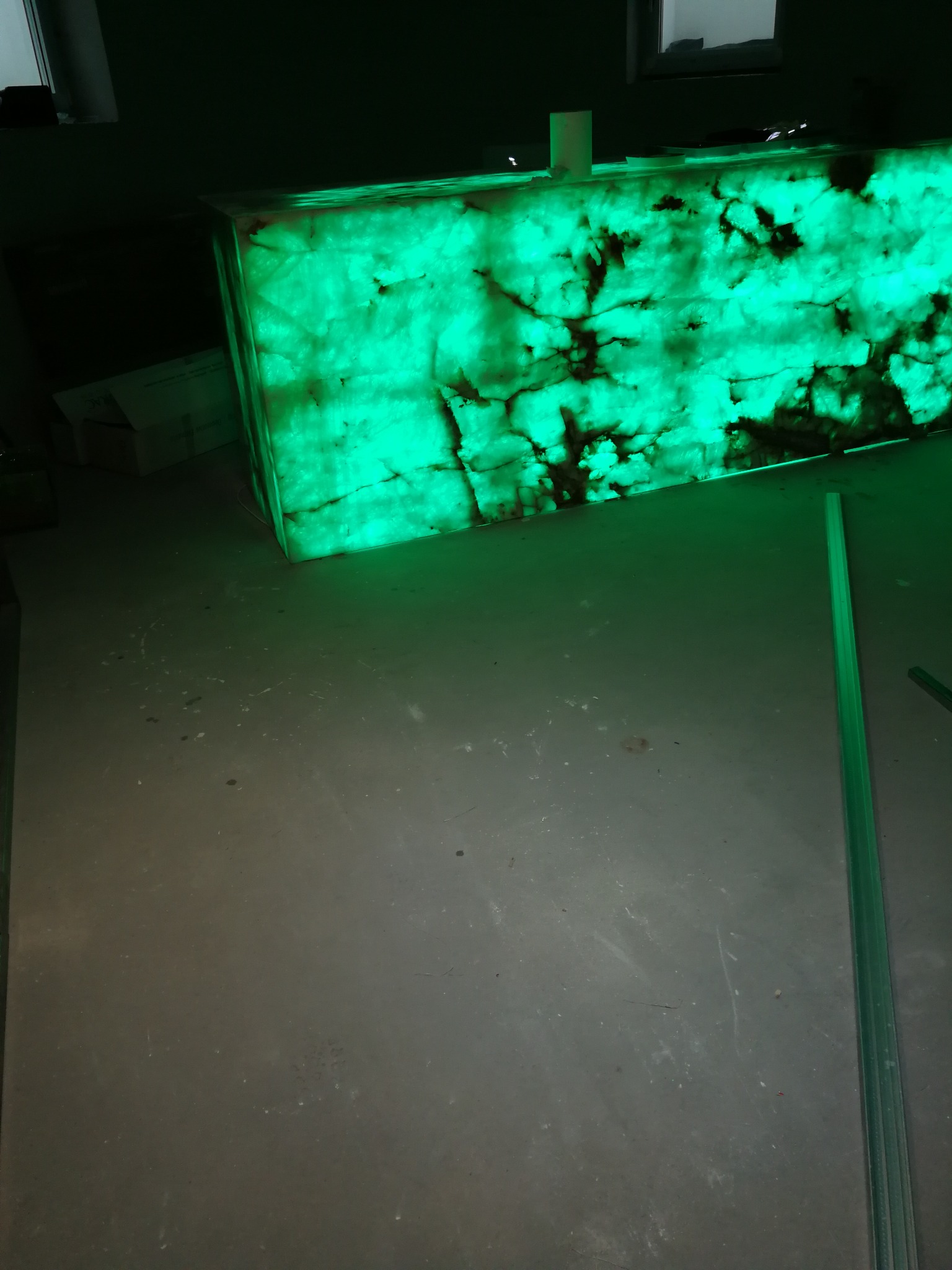 Podświetlony od spodu blat z onyksu w zielonej tonacji, widoczne ciemne żyły kamienia, surowy beton na podłodze, listwa przypodłogowa.