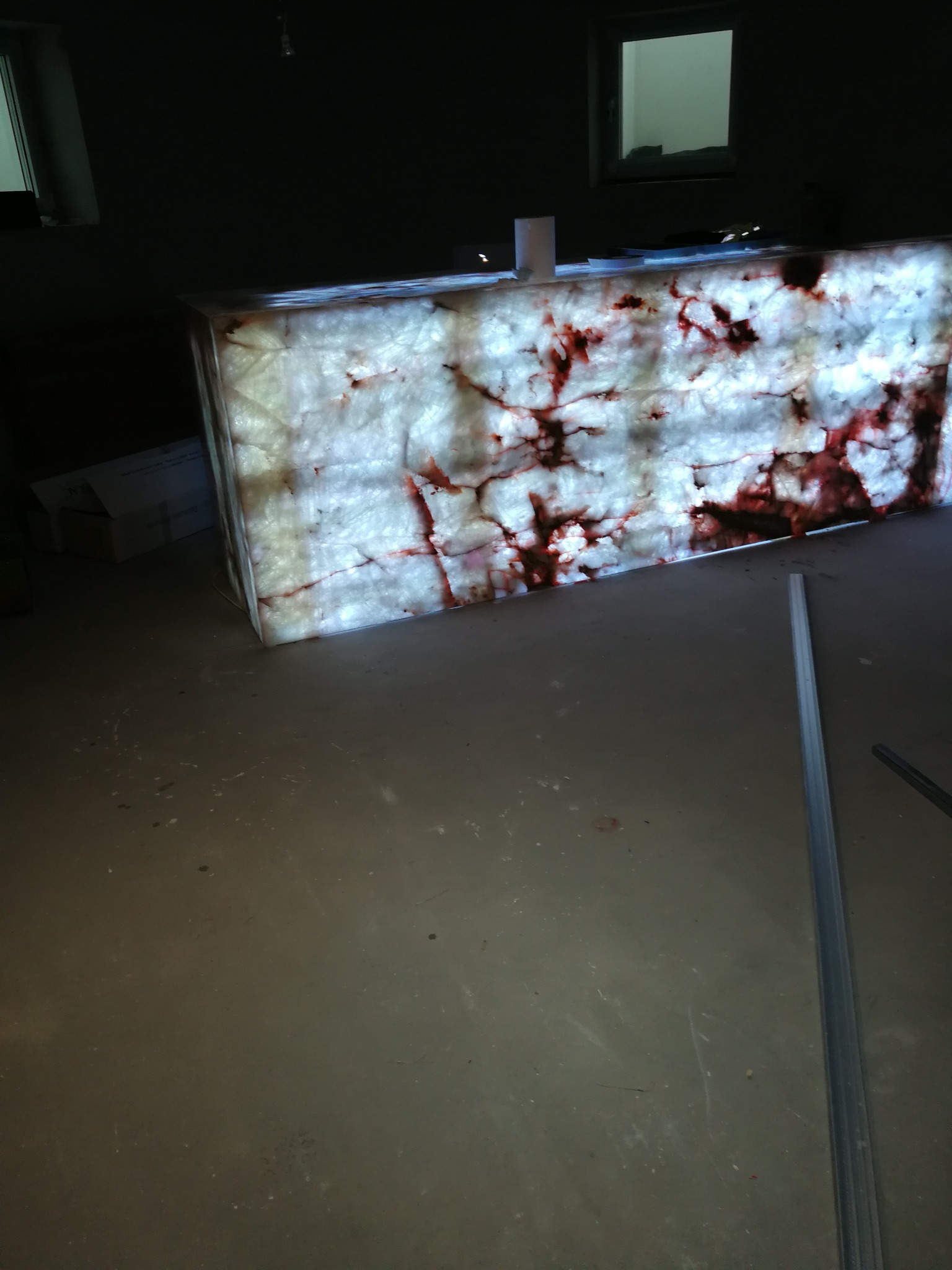 Podświetlony panel z onyksu z czerwonymi żyłkami, ustawiony w ciemnym pomieszczeniu, widoczne profile montażowe na podłodze.