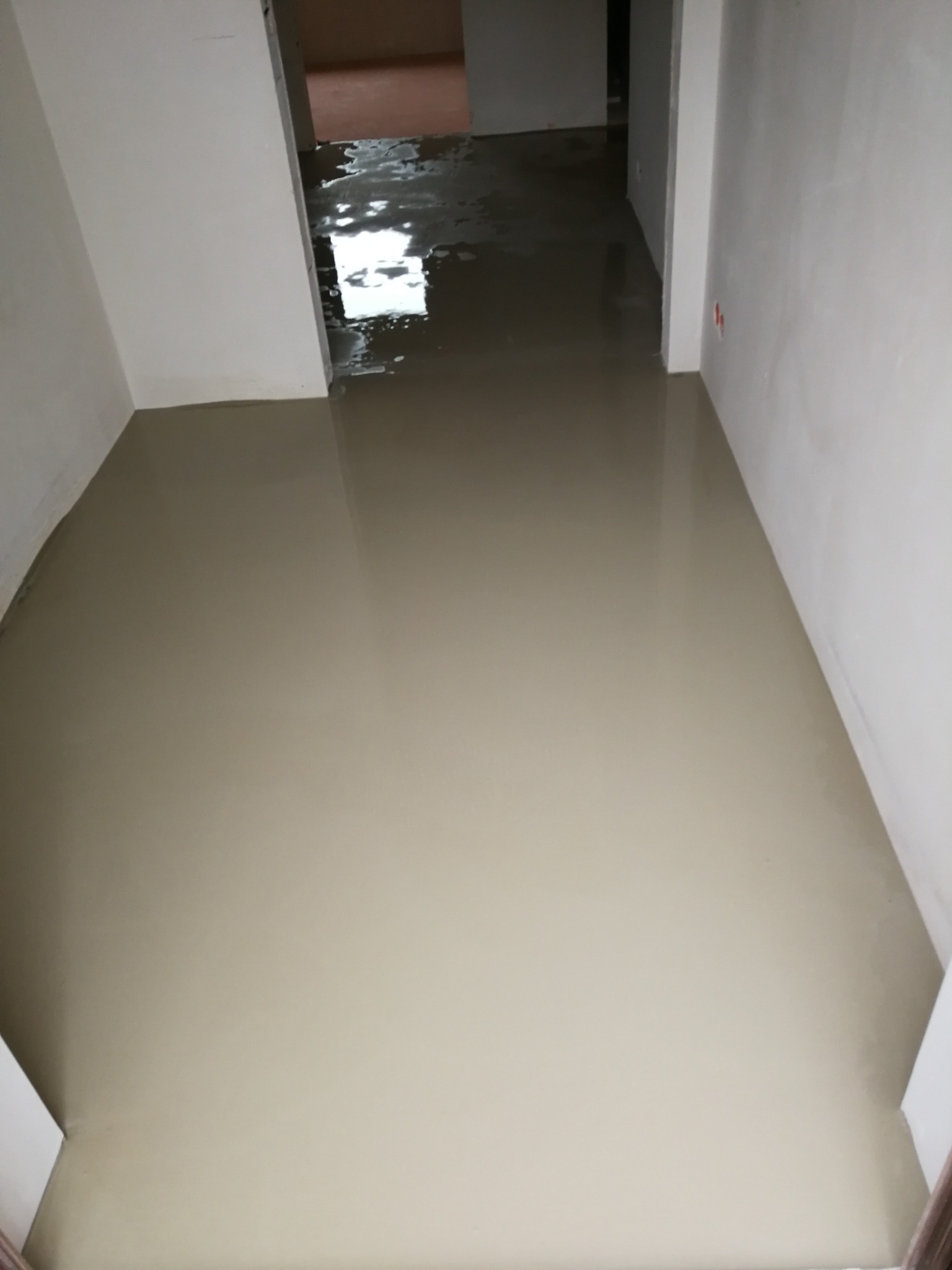 Świeżo wylana, gładka wylewka samopoziomująca na podłodze w trakcie remontu mieszkania, widoczna w perspektywie korytarza z białymi ścianami i odblaskami światła.