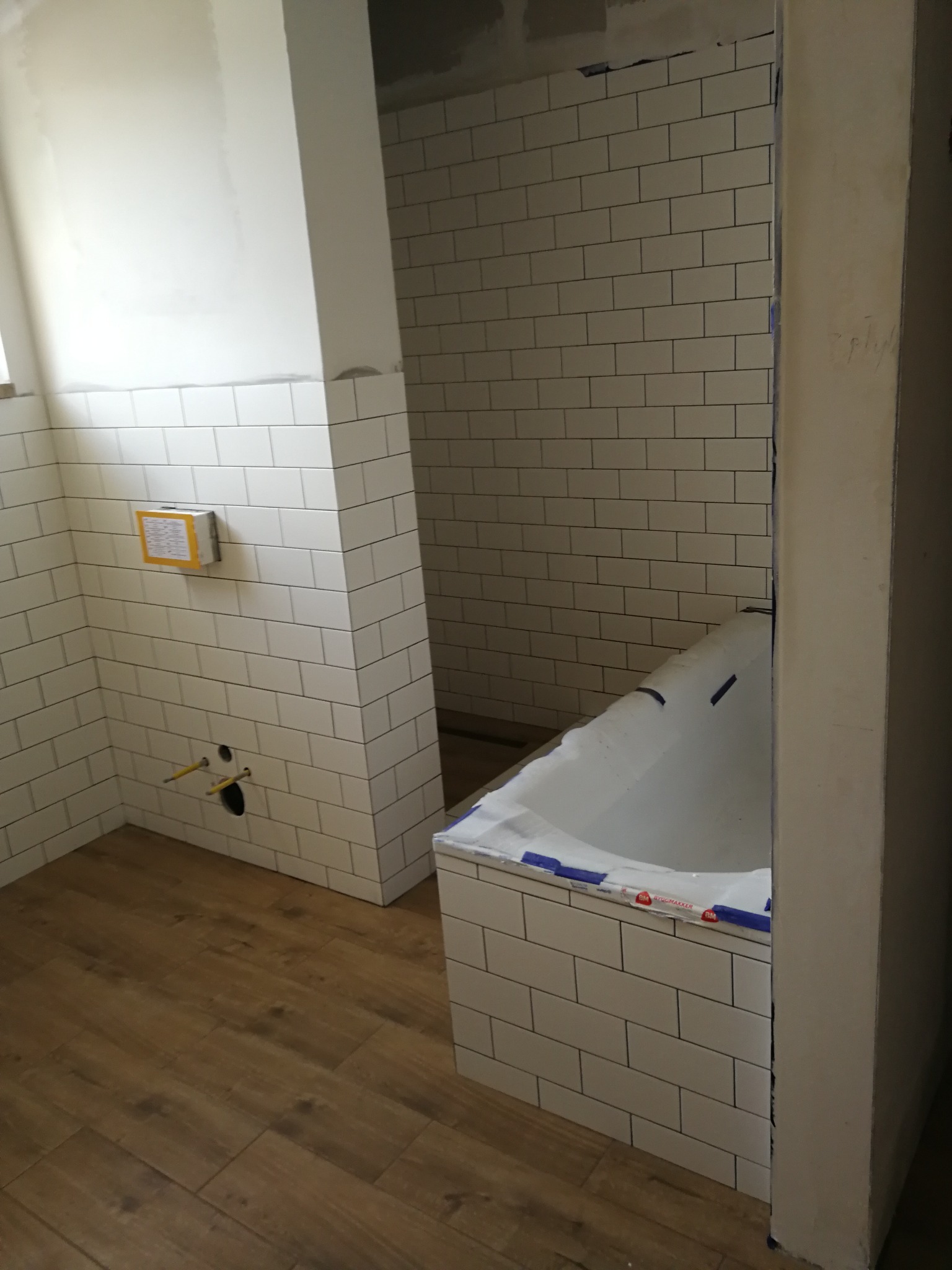 Wnętrze łazienki w trakcie wykańczania: białe, prostokątne płytki na ścianach, drewnopodobna podłoga, wanna zabezpieczona folią, widoczne instalacje hydrauliczne przed montażem armatury.