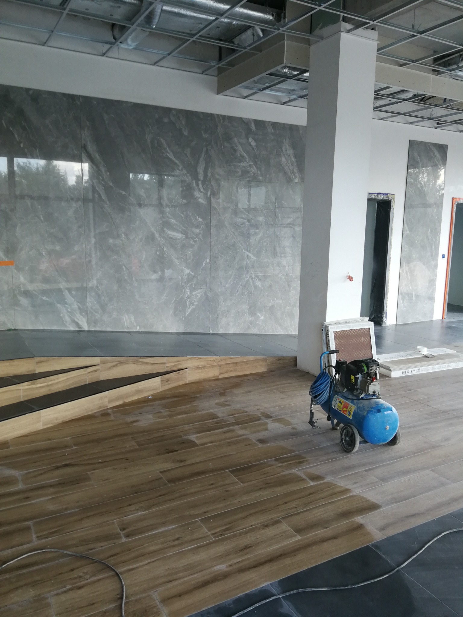 Wnętrze w trakcie prac wykończeniowych: ułożone płytki imitujące drewno na podłodze, szare marmurowe płytki na ścianie, widoczny kompresor i niezabudowany sufit podwieszany.