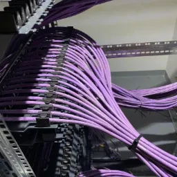 Równolegle ułożone, fioletowe kable sieciowe, uporządkowane w panelu krosowniczym, zabezpieczone czarnymi opaskami zaciskowymi, widoczne fragmenty metalowej konstrukcji szafy rack.