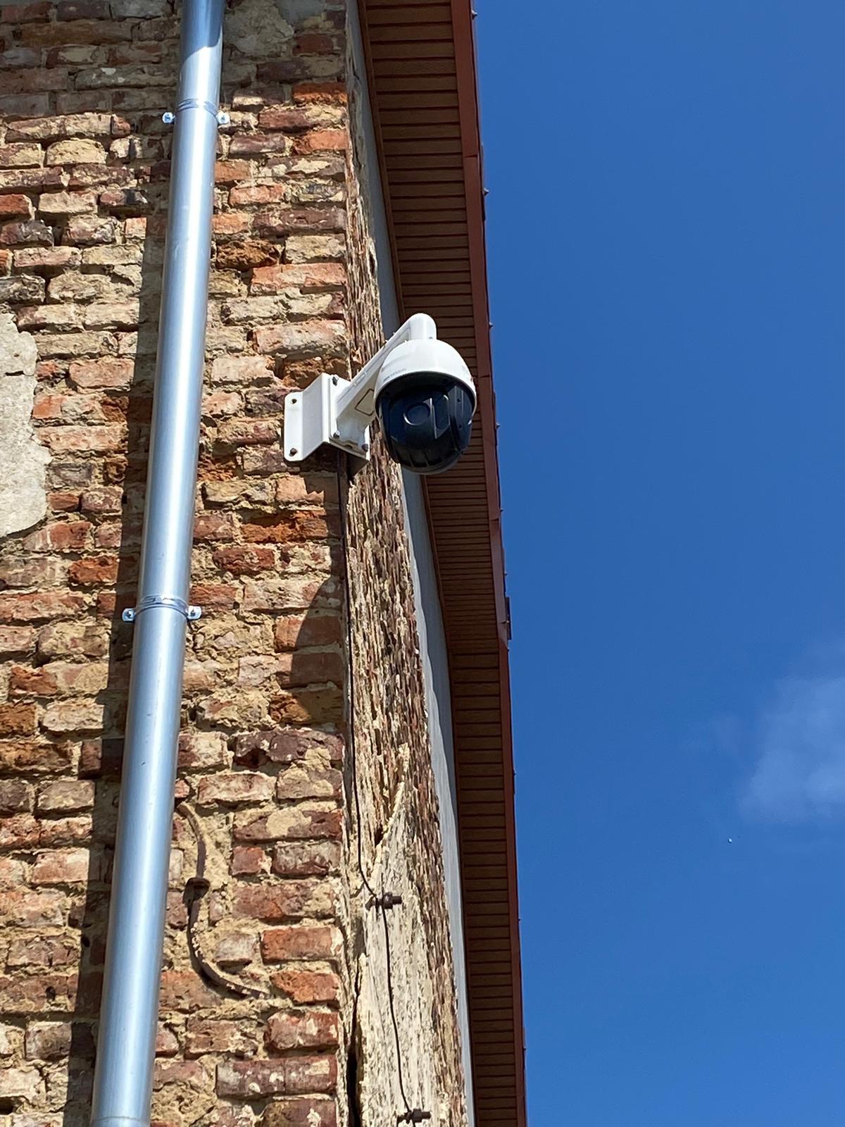 Kamera kopułkowa, biała na wsporniku, zamontowana na ceglanej ścianie budynku, z widoczną rynną spustową i fragmentem dachu na tle błękitnego nieba.
