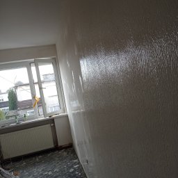 BadBud - Świeżo nałożona, mokra warstwa gładzi na ścianie w pokoju z oknem, widoczne wiaderka po farbie na podłodze zabezpieczonej folią.