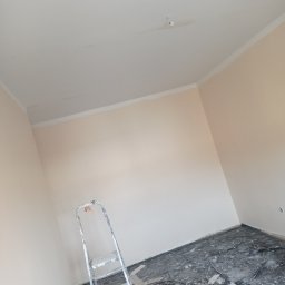 BadBud - Pokój po malowaniu ścian na jasny kolor z widoczną drabiną, zabezpieczoną podłogą folią malarską i torbą z narzędziami, ukazujący etap prac wykończeniowych.