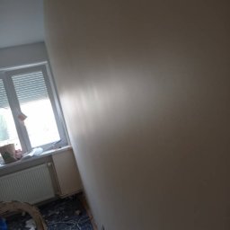 BadBud - Świeżo pomalowana ściana w odcieniach beżu, widoczne ślady wałka, okno z żaluzjami w tle, podłoga zabezpieczona folią, w rogu drabina malarska.