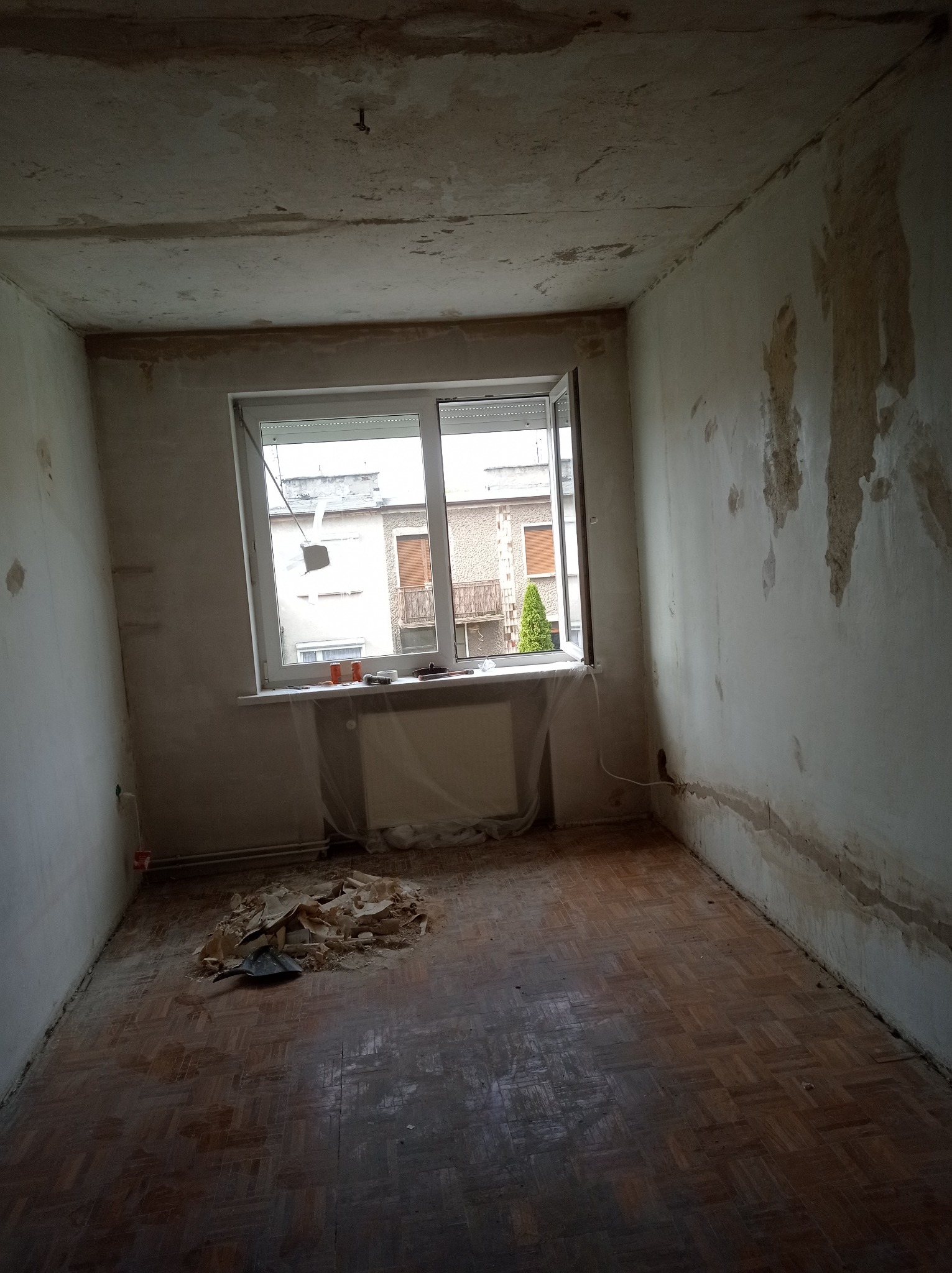 Pokój w trakcie przygotowań do remontu: ściany z uszkodzonym tynkiem, podłoga parkietowa, okno z widokiem na budynki, na podłodze sterta odpadów i szpachelka.