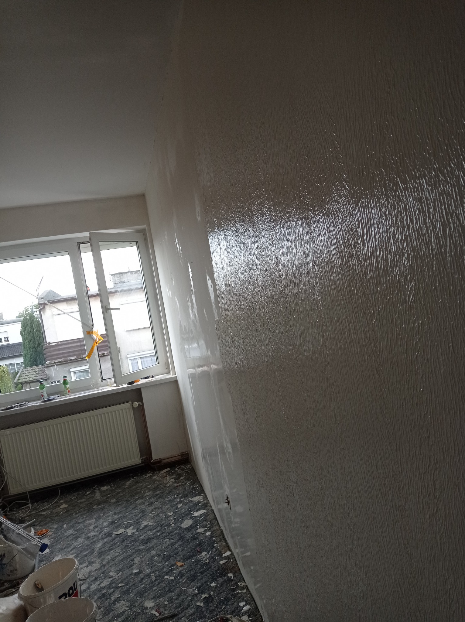 Świeżo nałożona, mokra warstwa gładzi na ścianie w pokoju z oknem, widoczne wiaderka po farbie na podłodze zabezpieczonej folią.