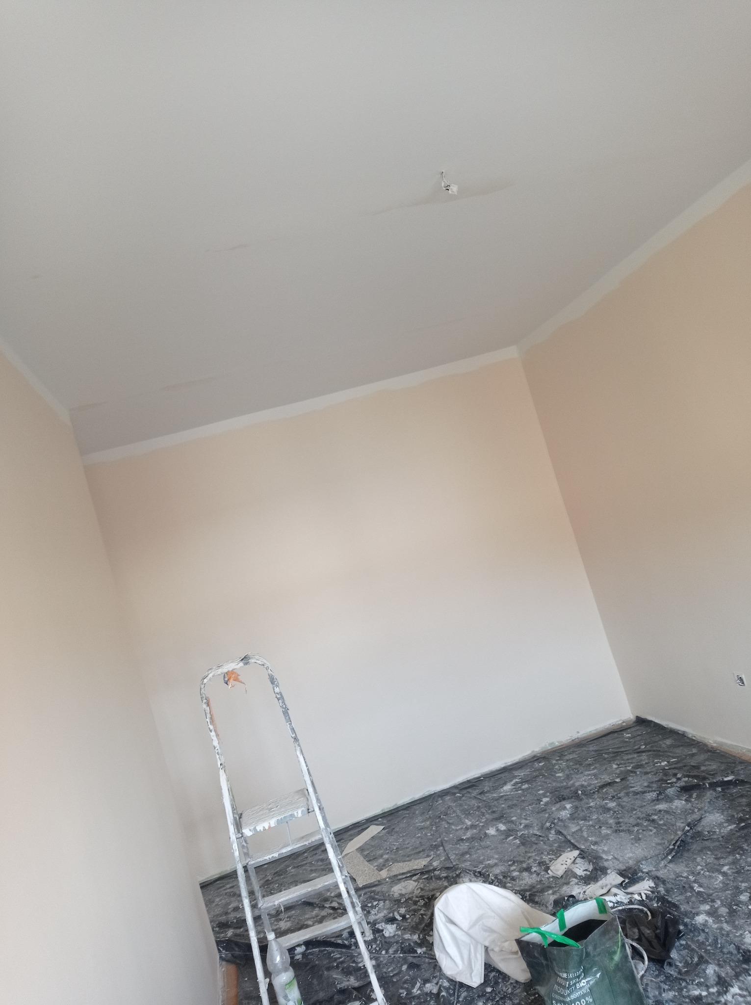 Pokój po malowaniu ścian na jasny kolor z widoczną drabiną, zabezpieczoną podłogą folią malarską i torbą z narzędziami, ukazujący etap prac wykończeniowych.