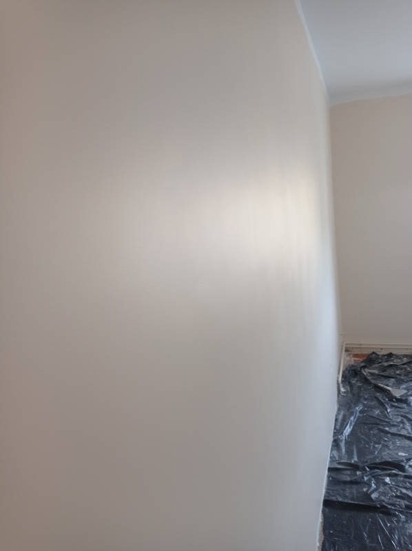 Świeżo pomalowana ściana w odcieniach beżu, widoczna listwa przypodłogowa i zabezpieczona podłoga folią malarską.