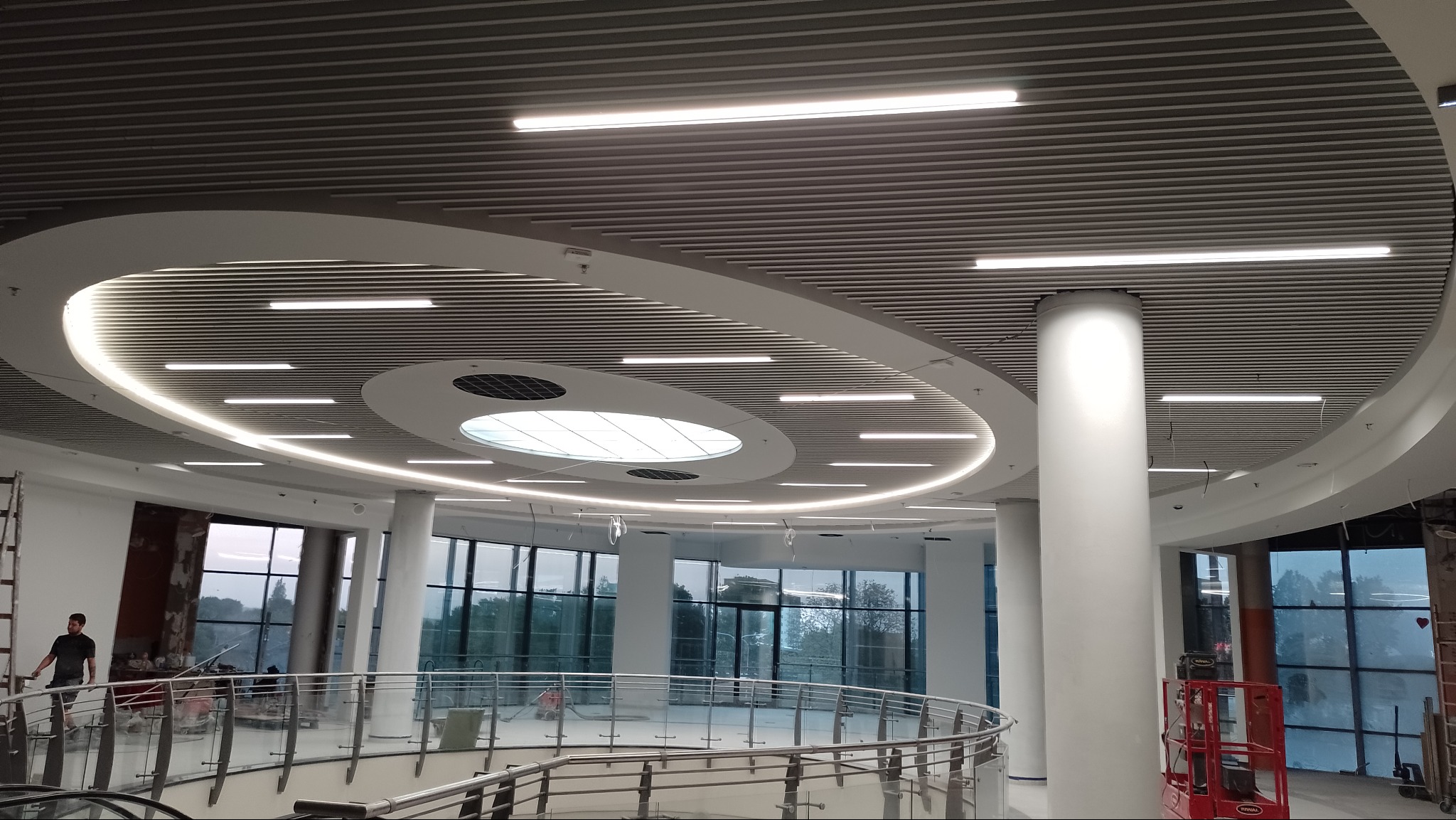 Nowoczesne oświetlenie sufitowe w przestronnym holu z kolumnami, widok na okna i balustradę, częściowo widoczne prace wykończeniowe.