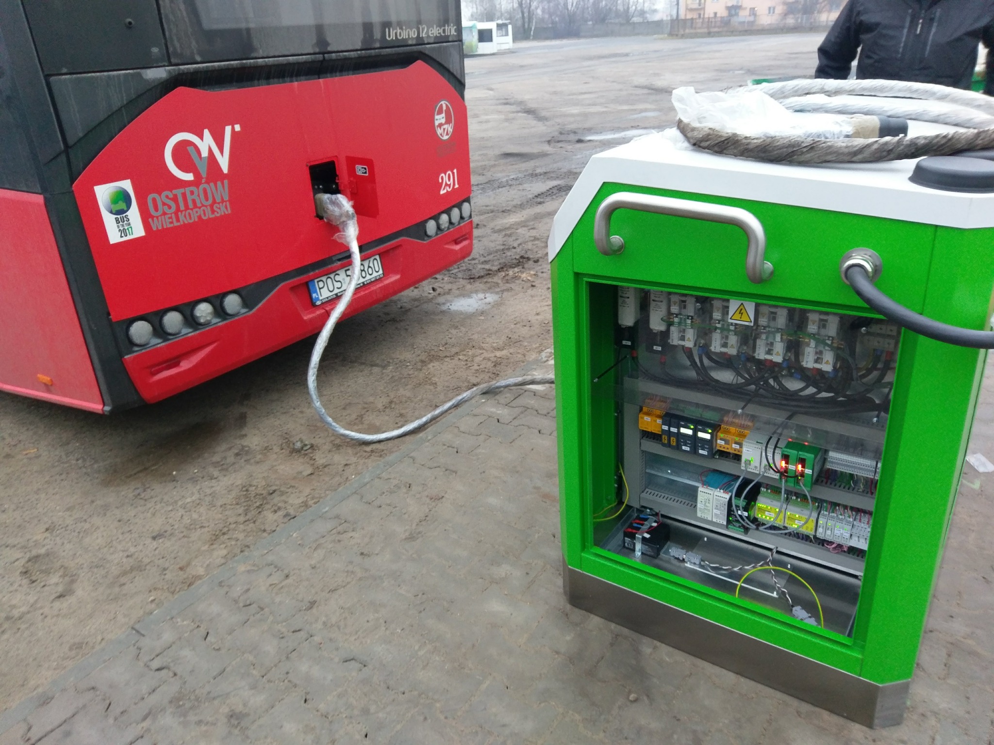 Czerwony autobus elektryczny marki Urbino 12 podczas ładowania z zielonej stacji ładowania na zewnątrz, widoczny kabel podłączony do gniazda autobusu.