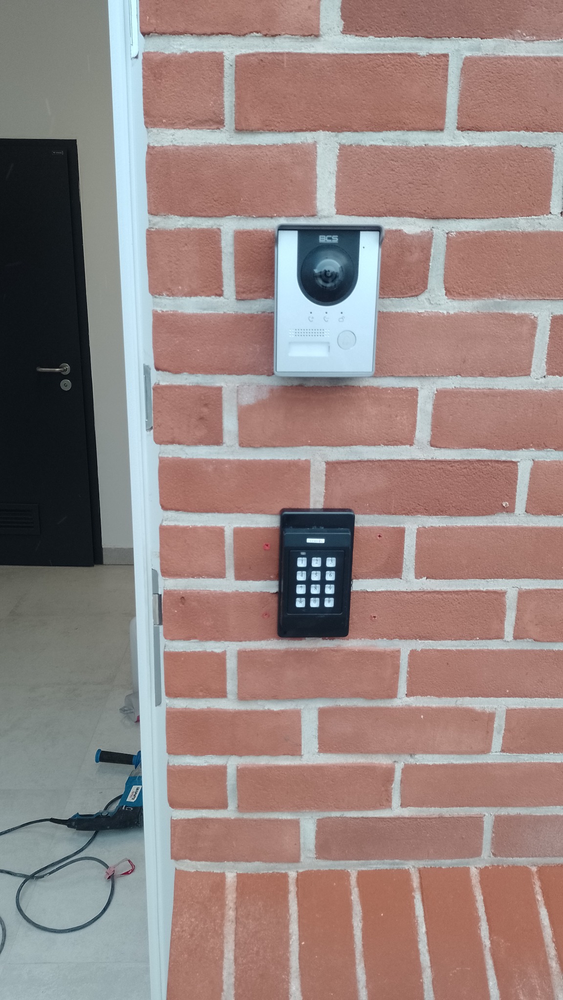 Zewnętrzny panel domofonowy z kamerą i klawiaturą numeryczną zamontowany na ceglanej ścianie, widoczny wiertarko-wkrętak akumulatorowy na podłodze.