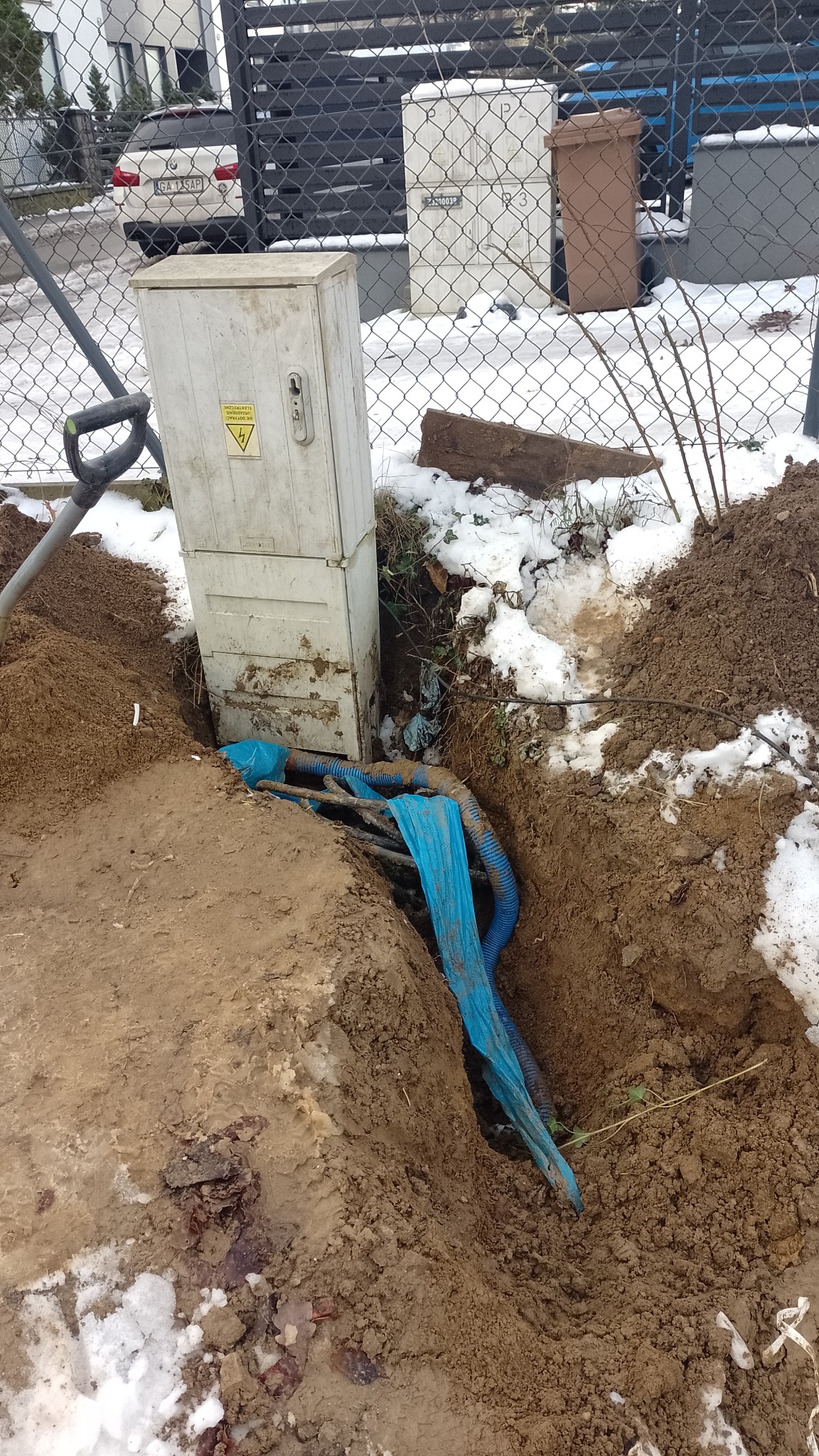 Odkopana skrzynka elektryczna na zewnątrz budynku, widoczne kable w niebieskiej osłonie w wykopie, resztki śniegu na ziemi.