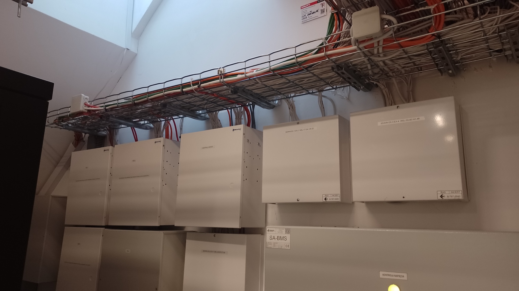 Rzędy białych skrzynek sterowniczych z widocznymi przewodami w metalowej korycie kablowej pod sufitem, etykiety informacyjne na obudowach.