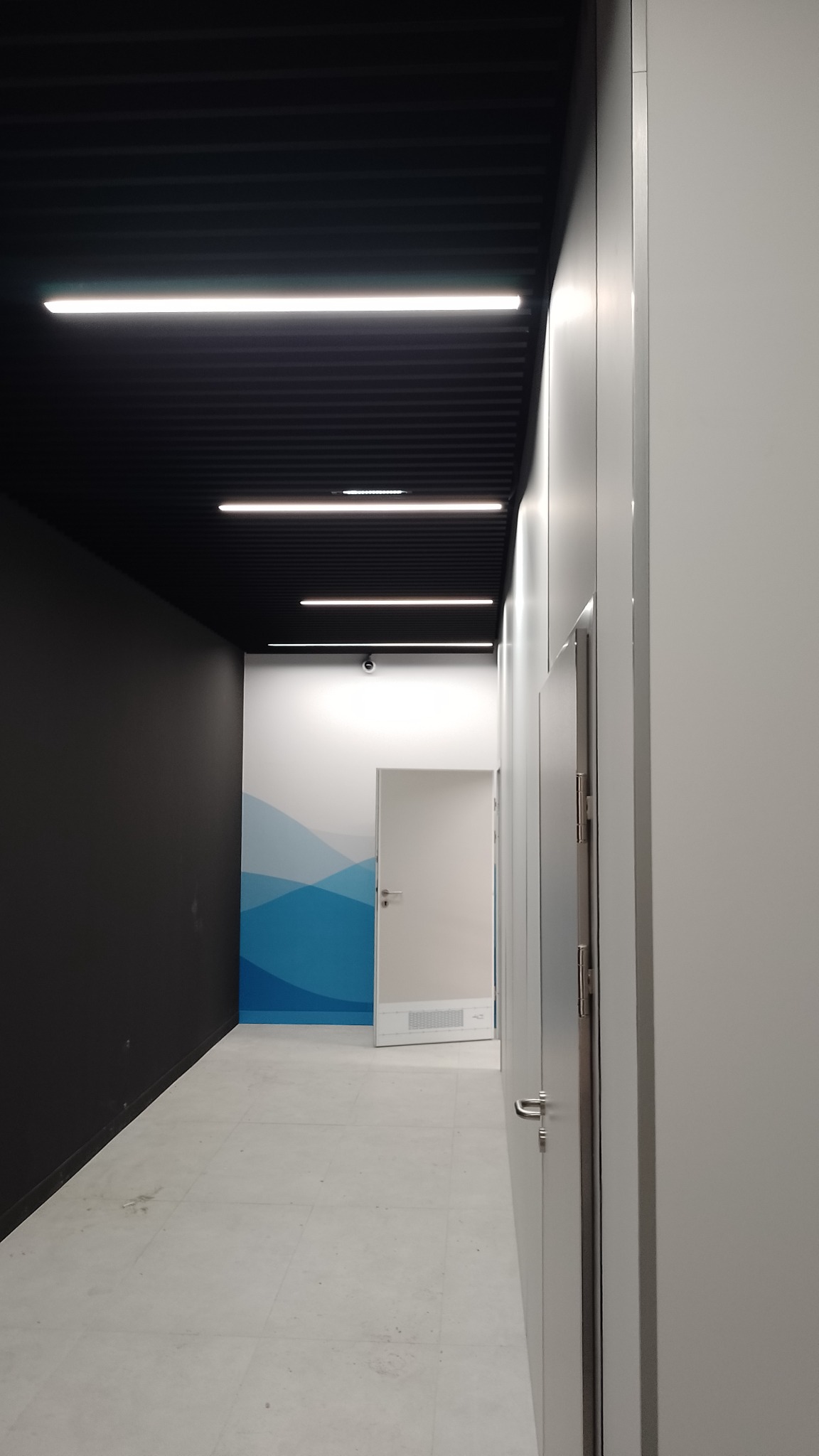 Korytarz biurowy z podwieszanym sufitem rastrowym i liniowymi oprawami oświetleniowymi LED, widoczna kamera monitoringu na ścianie.