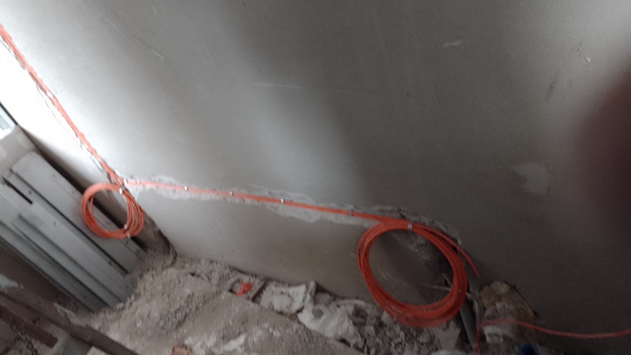 Pomarańczowy kabel elektryczny przymocowany do surowego, szarego sufitu i ściany w trakcie remontu, widoczne zwisające pętle kabla i gruz na podłodze.
