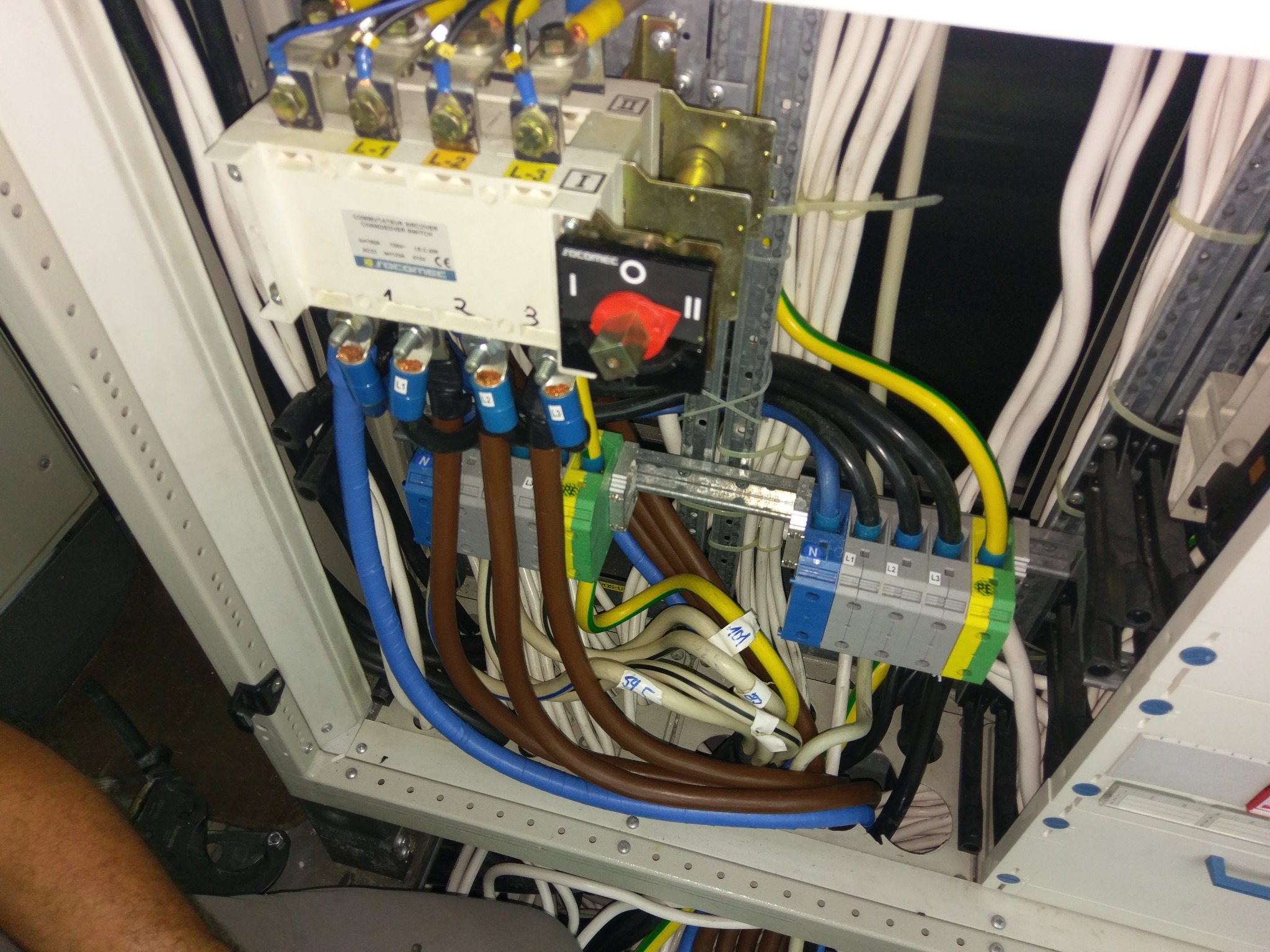 Rozdzielnica elektryczna z przełącznikiem, widoczne kable o różnych kolorach, oznaczenia na przewodach i elementach rozdzielnicy, wnętrze szafy sterowniczej z widoczną częścią buta osoby...