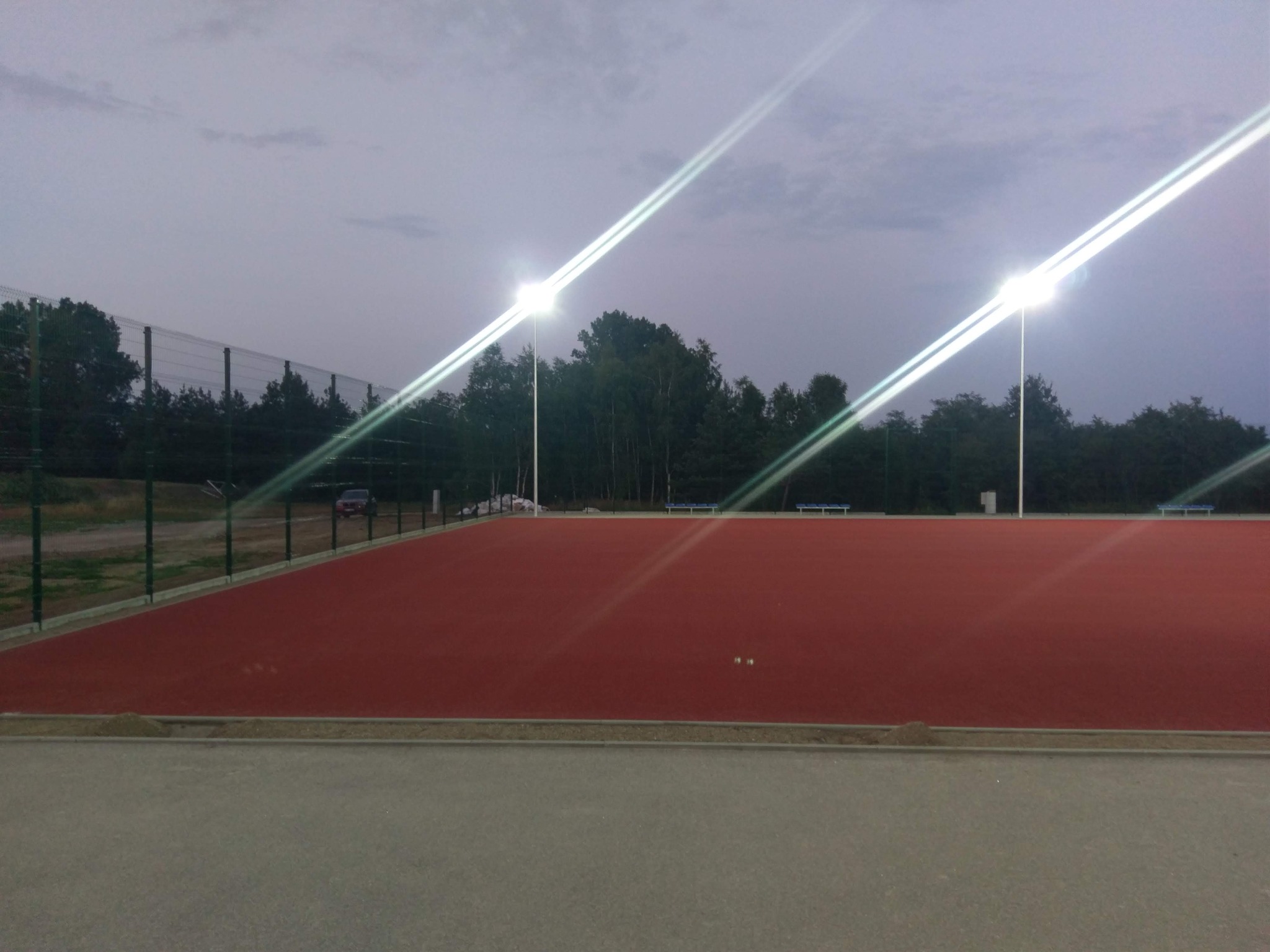 Oświetlony, czerwony kort sportowy z siatką ochronną i drzewami w tle, sfotografowany o zmierzchu.