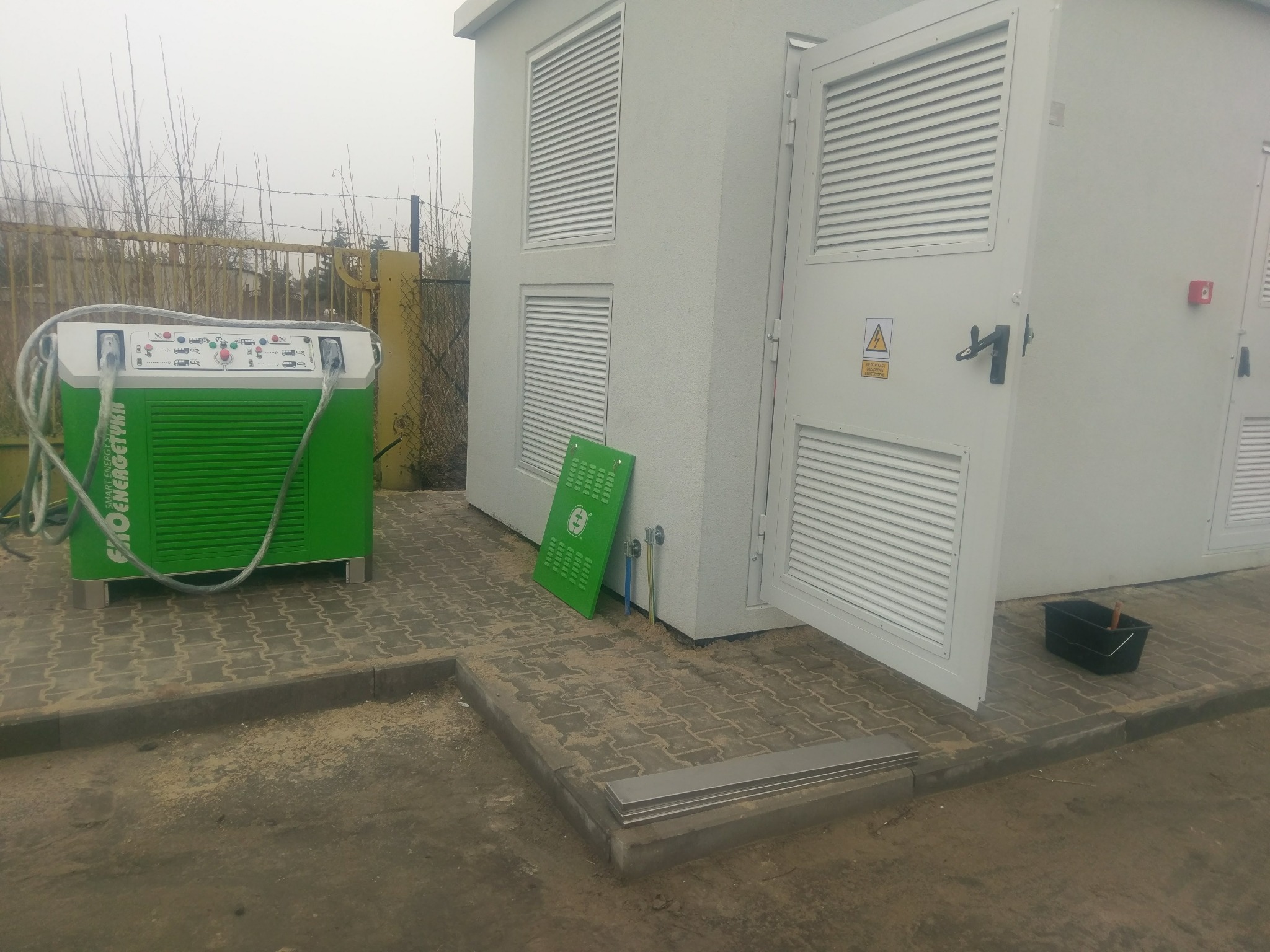 Zewnętrzna stacja transformatorowa z otwartymi drzwiami, zielona przenośna jednostka pomiarowa z przewodami oraz skrzynka narzędziowa obok wejścia.