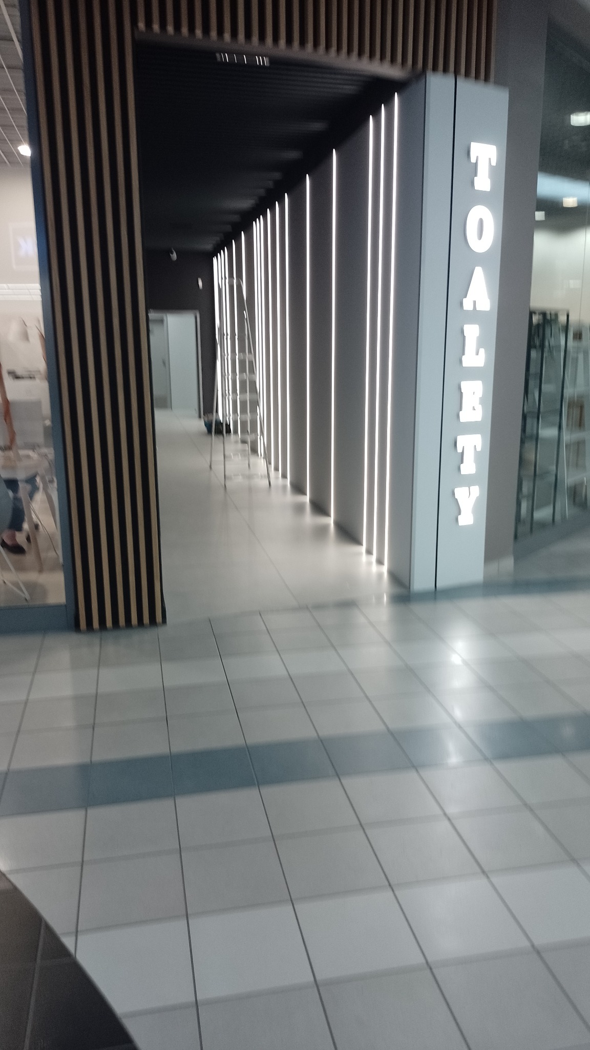 Oświetlenie LED w korytarzu publicznym z napisem 'TOALETY' na boku, widoczna drabina w tle, kafelki na podłodze.