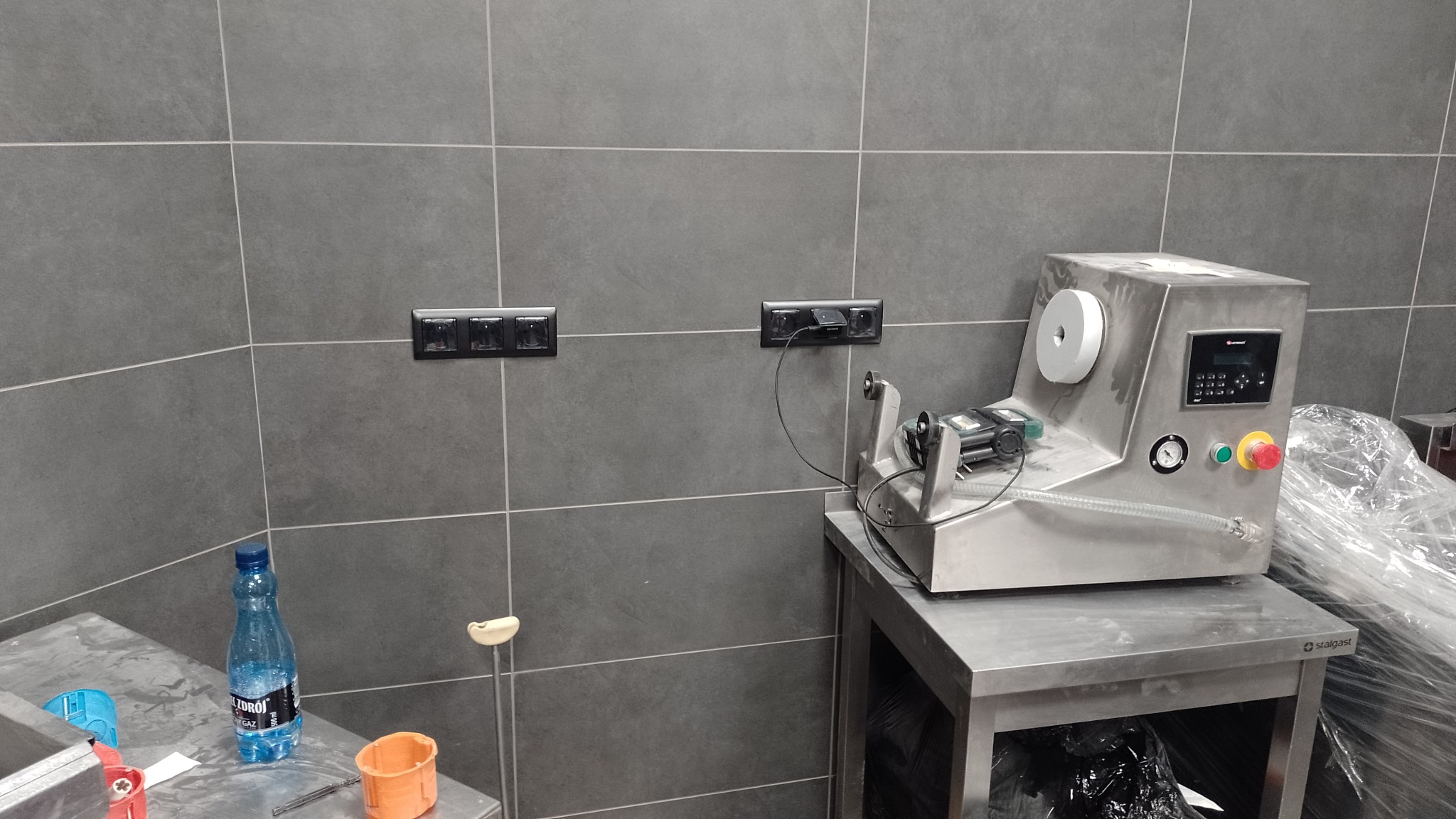 Szare płytki ścienne z zamontowanymi gniazdkami elektrycznymi, do jednego z nich podłączone jest urządzenie stojące na metalowym stole. Na stole widoczna butelka wody i pomarańczowy pojemnik.