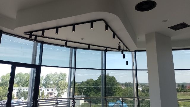 Wnętrze biurowe z nowoczesnym systemem oświetlenia szynowego z reflektorami punktowymi, widok na zewnątrz przez duże okna z czarnymi ramami.