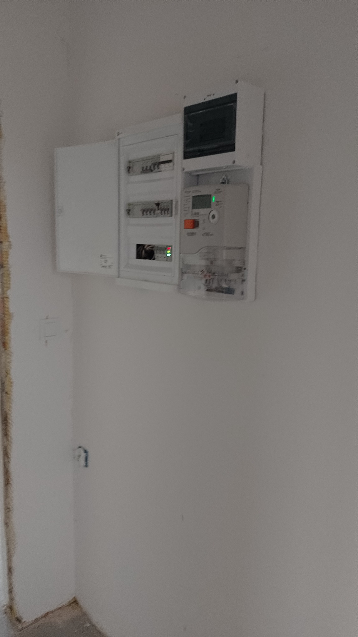 Rozdzielnica elektryczna z otwartymi drzwiczkami, widoczny licznik z zieloną diodą, instalacja w surowym stanie, ściany niepomalowane.