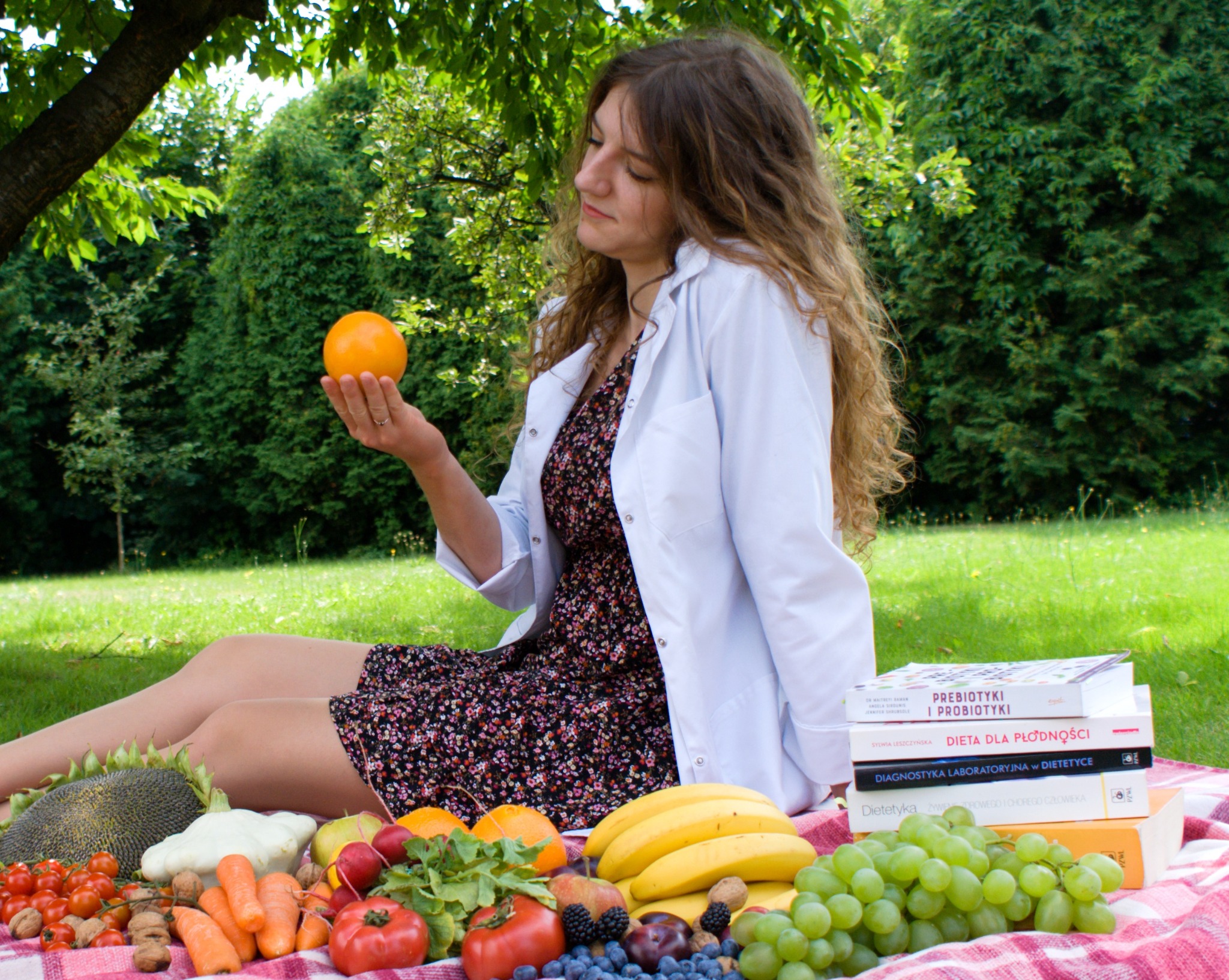 Kobieta w białym fartuchu siedzi na kocu piknikowym na trawie, trzymając pomarańczę w dłoni, obok leżą warzywa, owoce oraz stos książek o tematyce żywieniowej i dietetyce.