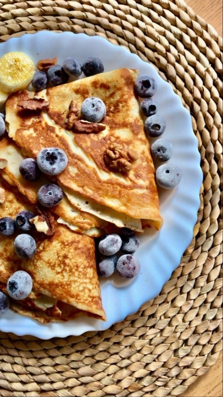 Złociste naleśniki na białym talerzu, udekorowane mrożonymi borówkami, orzechami włoskimi i plasterkiem banana, umieszczone na plecionej podkładce.