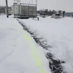 Ścieżka odśnieżona na dachu przemysłowym, z widocznymi urządzeniami klimatyzacyjnymi i wentylacyjnymi, oznaczona jaskrawym, zielonym kolorem, w tle drzewa bez liści i szare niebo.