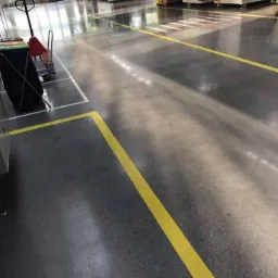 Błyszcząca, szara posadzka przemysłowa z wyraźnie odznaczonymi żółtymi liniami wyznaczającymi ciągi komunikacyjne, w tle widoczny wózek transportowy i regały magazynowe.