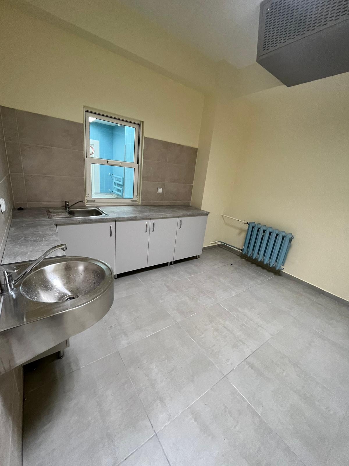 Pusta umywalnia z nierdzewnej stali, szare płytki podłogowe i ścienne, niebieski grzejnik żeberkowy, białe szafki pod oknem, widok na inne pomieszczenie przez okno.