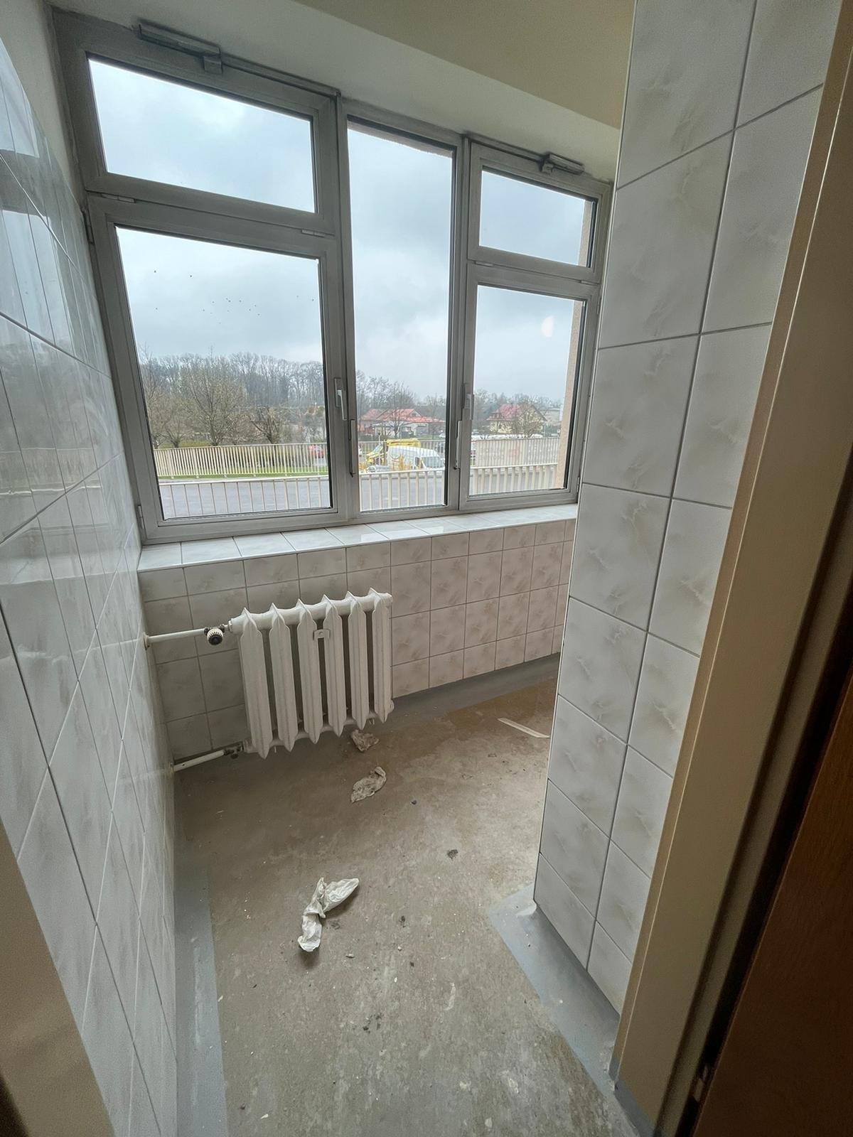 Balkon z oknami i grzejnikiem przed czyszczeniem, widoczne pozostałości po remoncie, jasne płytki na ścianach i podłodze, okno z widokiem na zewnątrz.