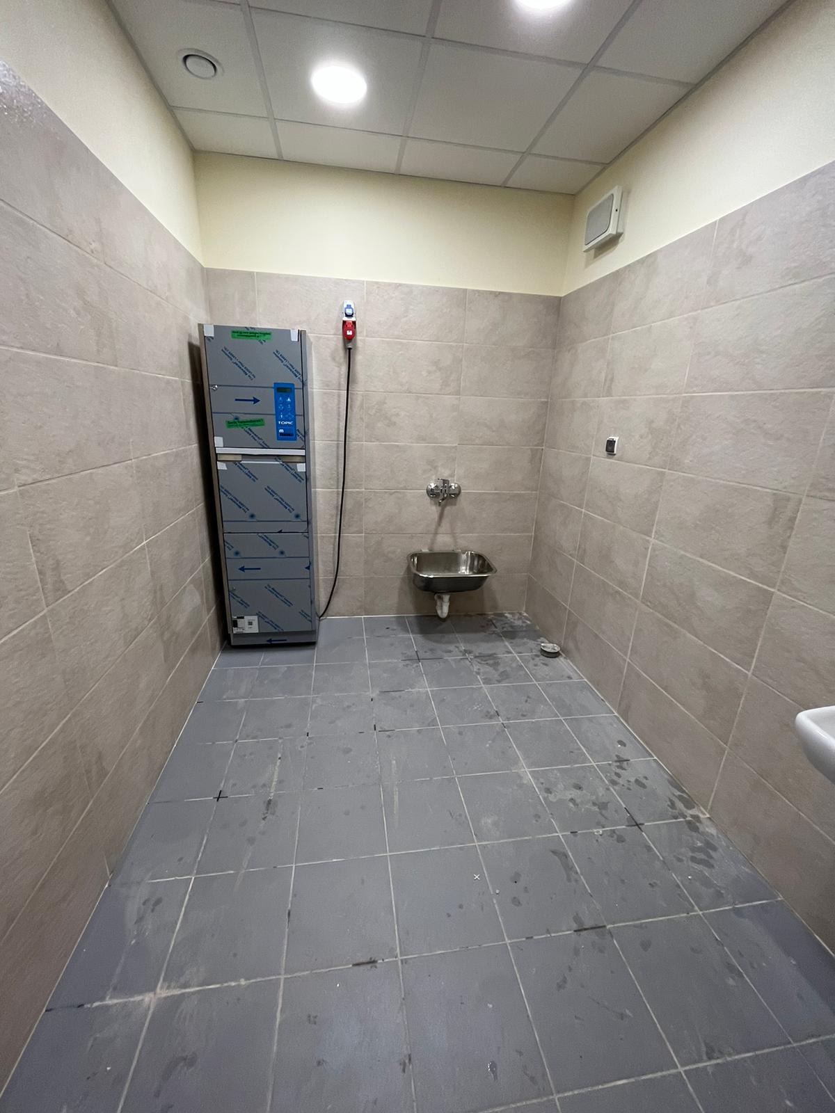 Wnętrze umywalni z szarą podłogą w płytkach, beżowymi ścianami, metalową szafką na środki czystości i umywalką ze stali nierdzewnej, widoczne ślady po czyszczeniu podłogi.
