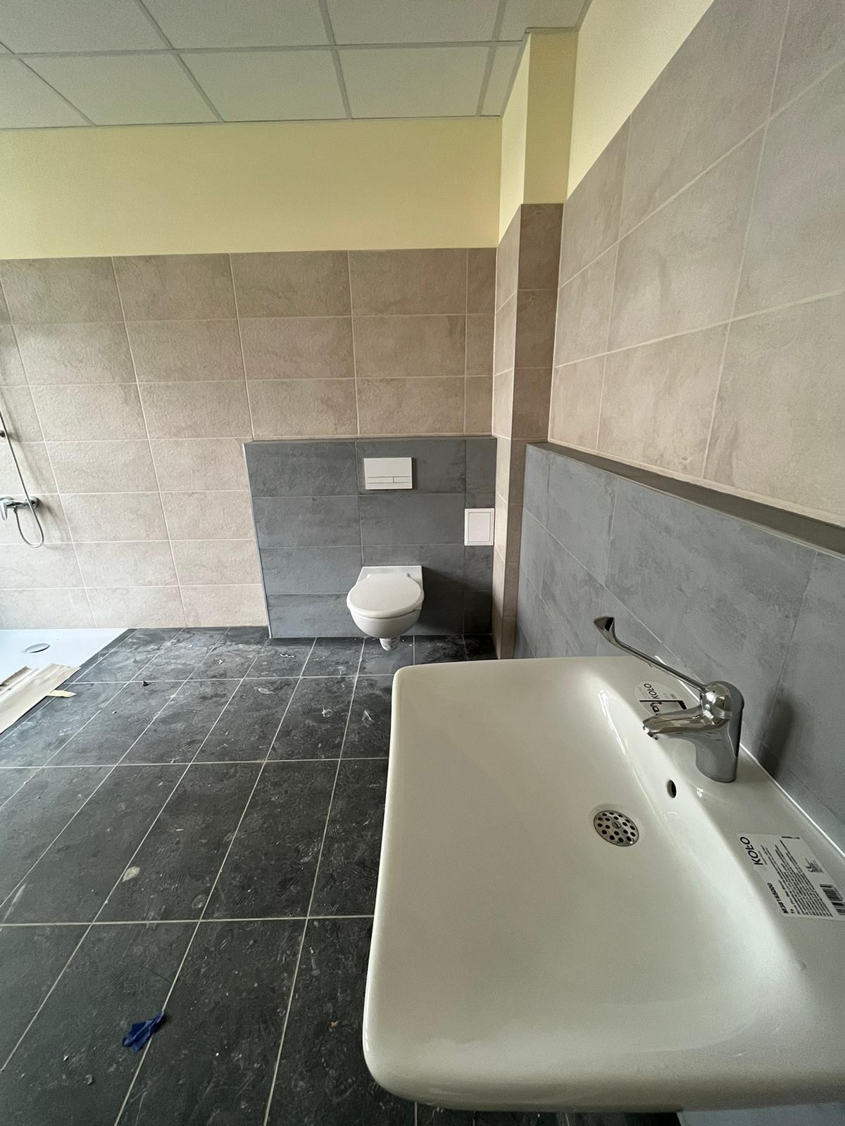 Wnętrze łazienki w trakcie remontu: widoczna umywalka marki Koło, toaleta podwieszana i częściowo wykończone płytki ścienne w odcieniach szarości i beżu, na podłodze ciemne płytki w trakcie...