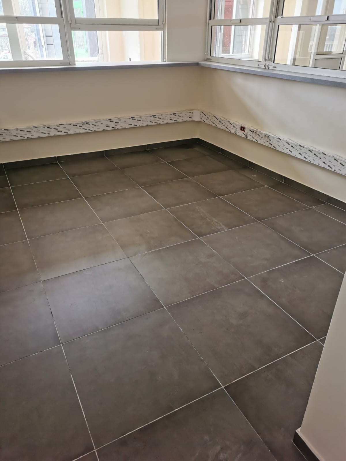 Puste pomieszczenie z szarymi płytkami na podłodze, listwami przypodłogowymi i osłonami instalacji elektrycznej w folii ochronnej, przygotowane do użytku.